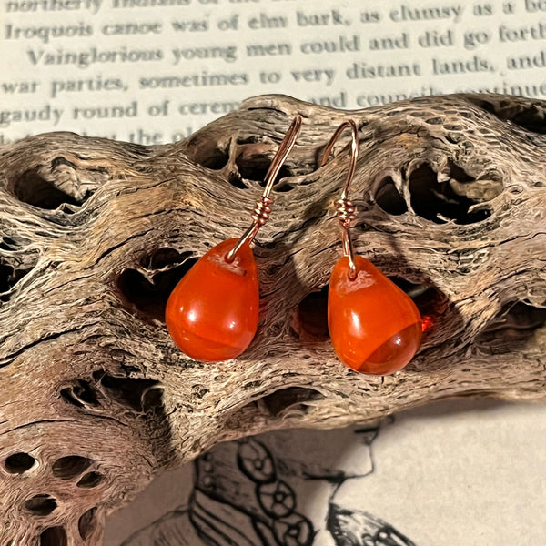 Pair of orange earrings on a piece of driftwood with text and butterfly illustration in the background